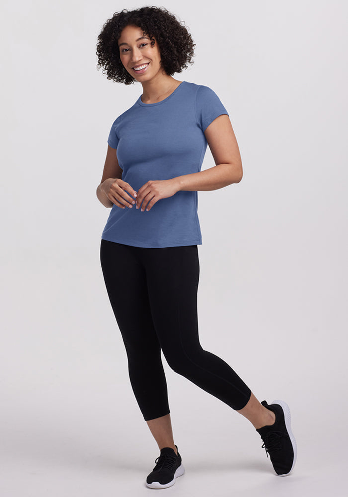 A woman with curly hair smiles while wearing the Woolx Addie Crew Neck – Skyline, black leggings, and black sneakers against a plain light background.