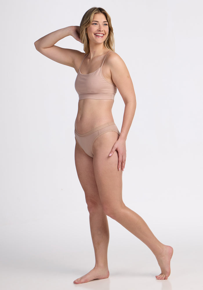 A woman stands barefoot on one leg, smiling with her left hand on her head, wearing a light beige bra and Woolx Roxie Bikini Underwear in Morning Latte against a plain white background.