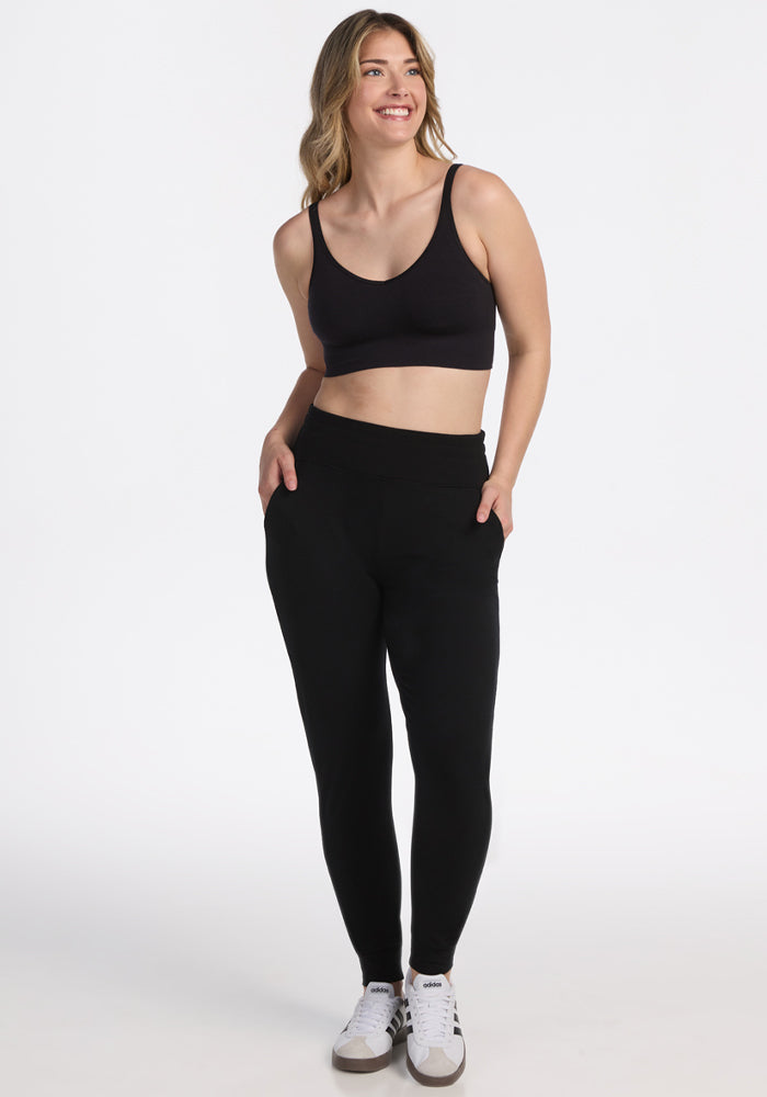 A woman smiles with her hands in the pockets of Woolx Madden Joggers - Black, paired with a black sports bra and white sneakers, against a plain white background. 