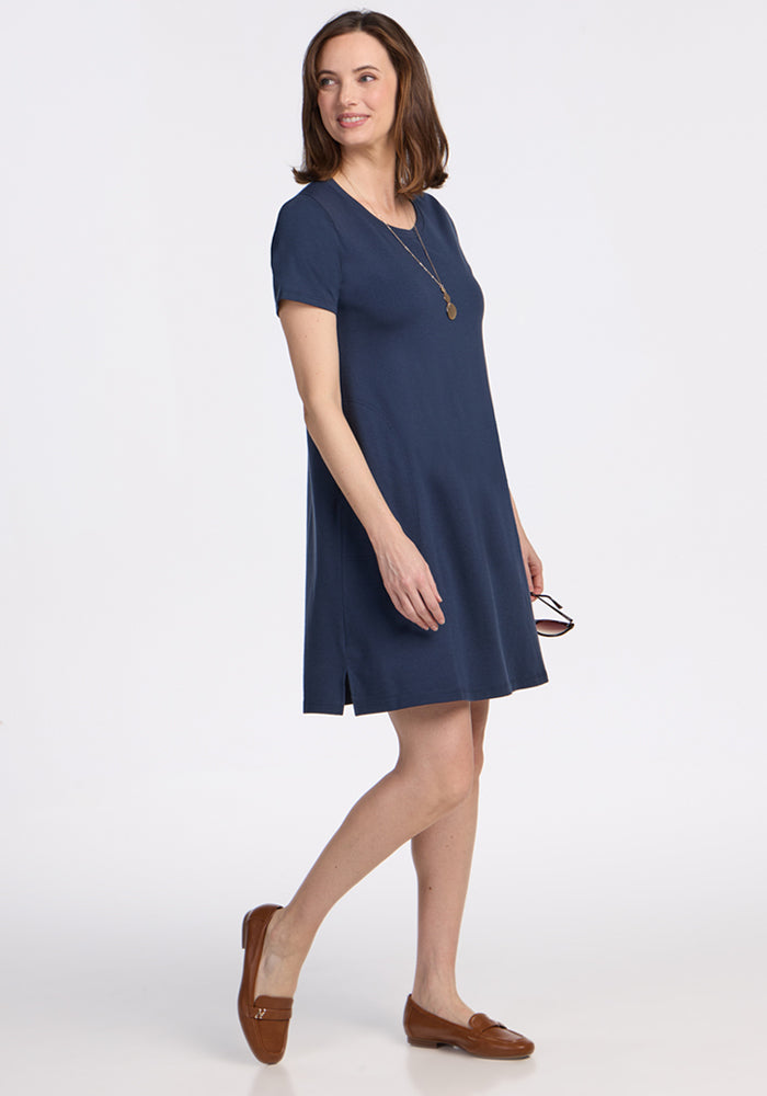 A woman smiles against a white background in the Woolx Georgie Dress – Deep Navy, a short-sleeved, knee-length Merino wool swing dress, styled with brown loafers, a necklace, and sunglasses in hand.