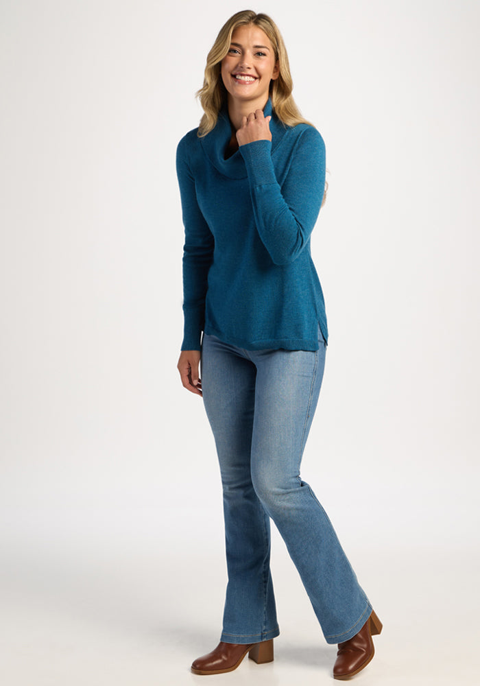 Woman in Woolx Josie Cowl Neck Sweater - Blue Pine and jeans, smiling with hand near neck, wearing brown boots.