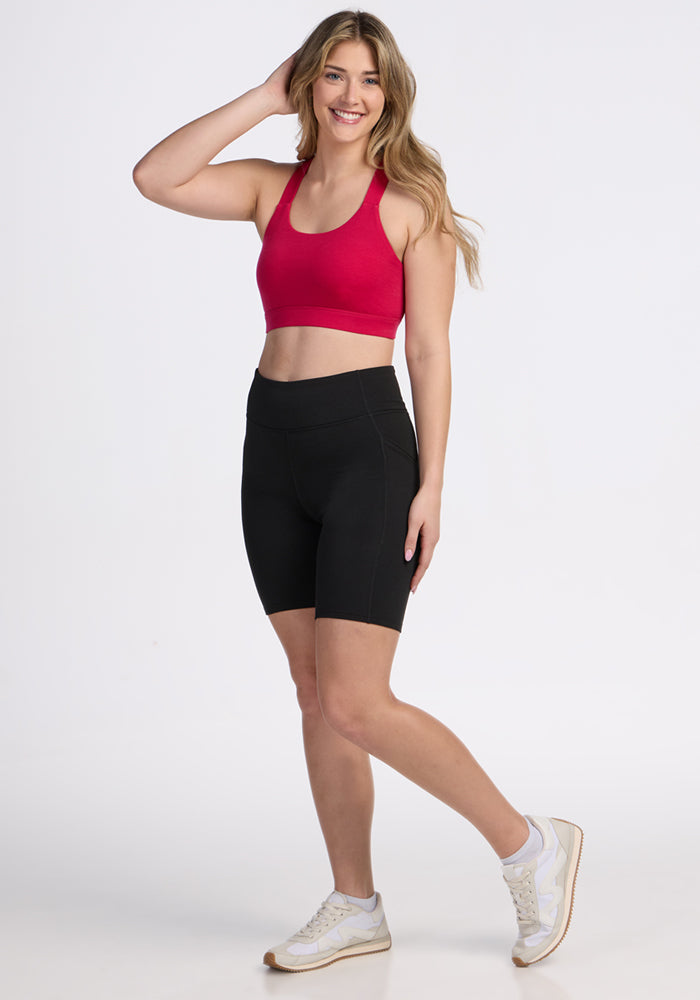 A woman with long, wavy blonde hair smiles while posing in the Woolx Livi Bra - Poppy Spark, black high-waisted bike shorts, and white sneakers against a plain white background.