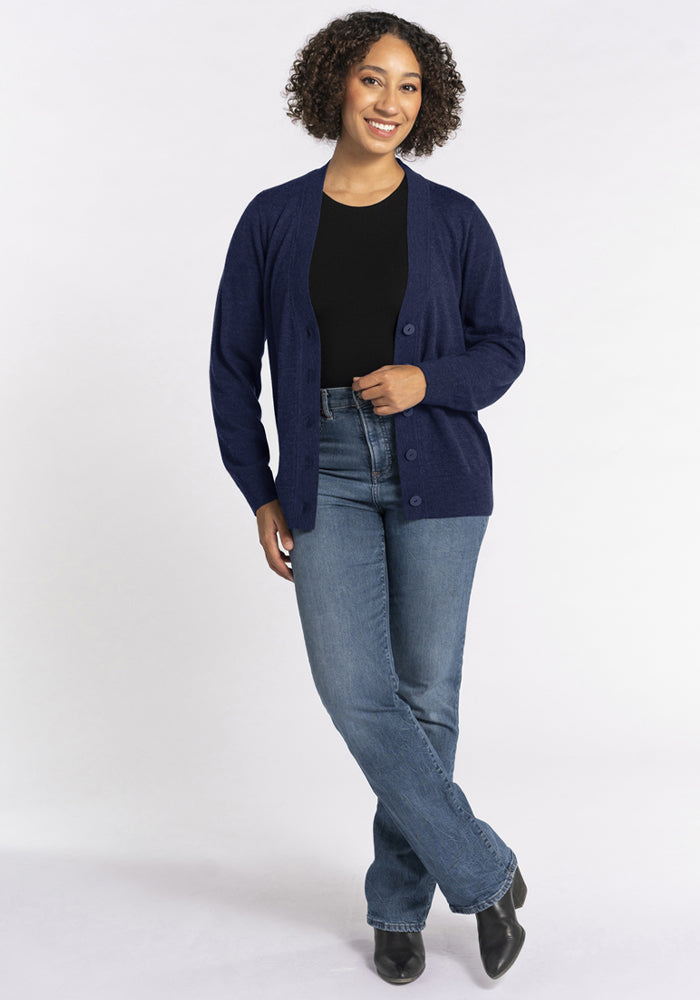 Smiling woman in Woolx Gia Sweater - Ocean Cavern, black top, blue jeans, and boots stands against a white background.