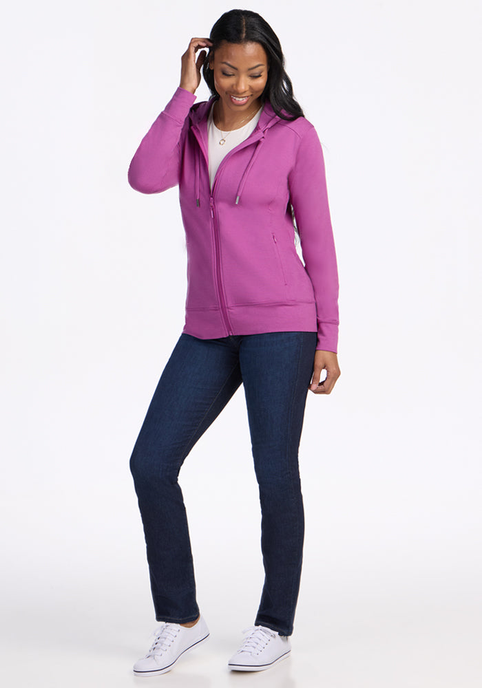 A woman smiles in the Woolx Ryann Hoodie - Purple Wine, paired with a white shirt, dark blue jeans, and white sneakers against a plain white background. She touches her hair with one hand and appears relaxed.