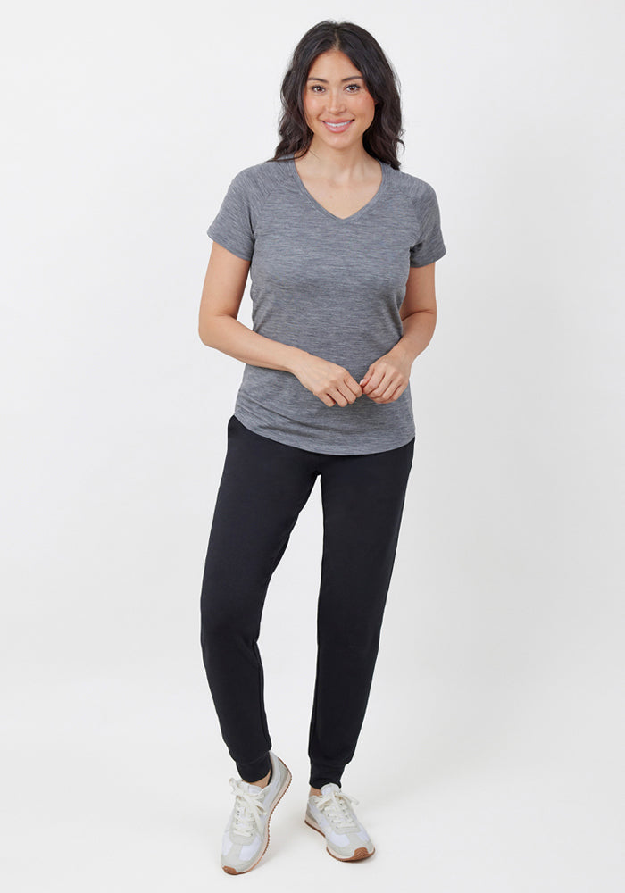 A woman with long dark hair smiles, wearing Woolx Madden Joggers in black, a gray V-neck T-shirt, and white sneakers against a white background.