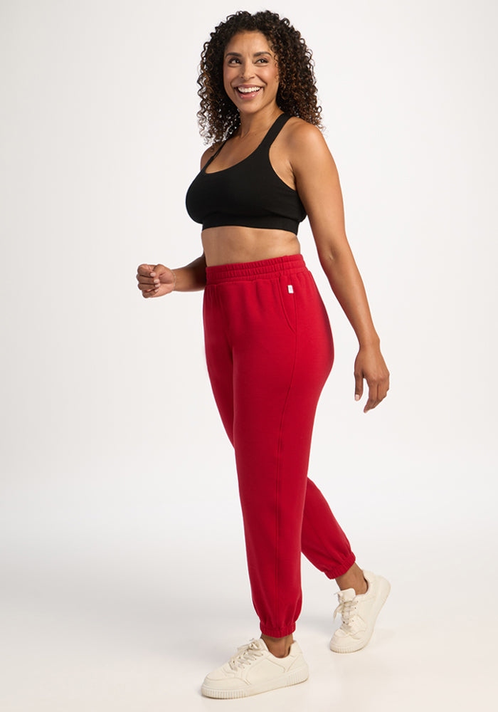 Woman in Woolx Parker Sweatpants - Maraschino Cherry, black sports bra, and white sneakers smiles in a studio.