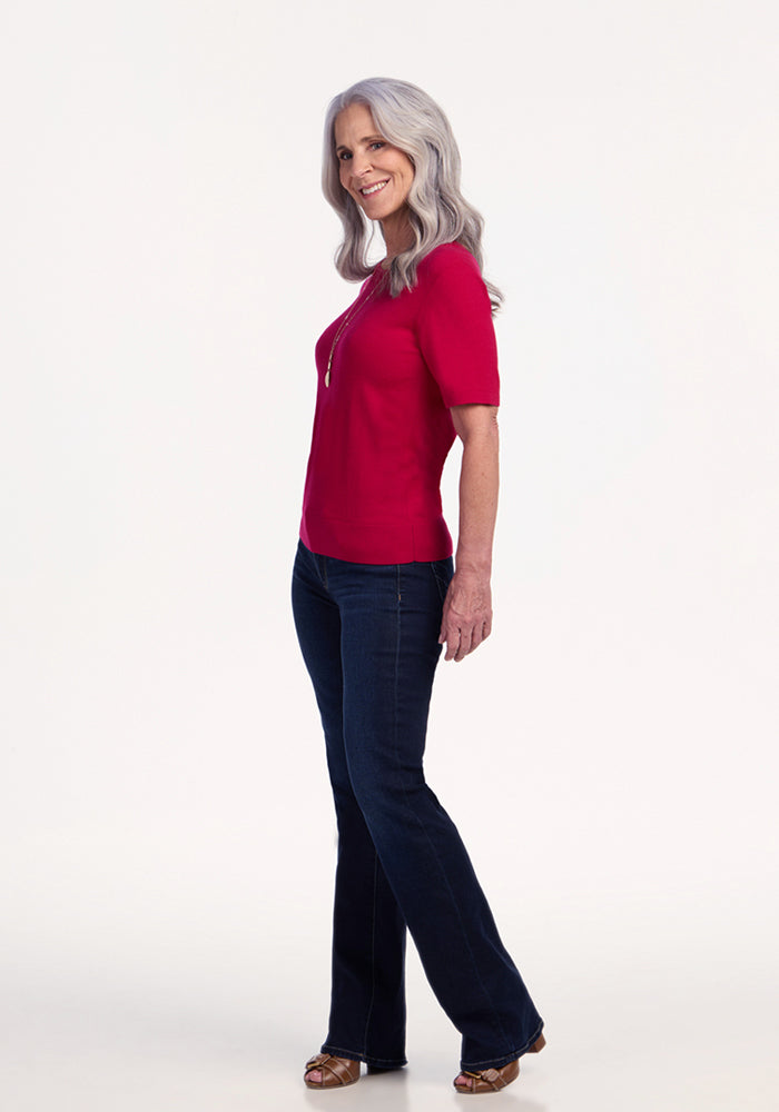A smiling older woman with long gray hair stands sideways against a plain white background, wearing the Woolx Anna Crew Sweater in Poppy Spark, paired with dark blue jeans and sandals.