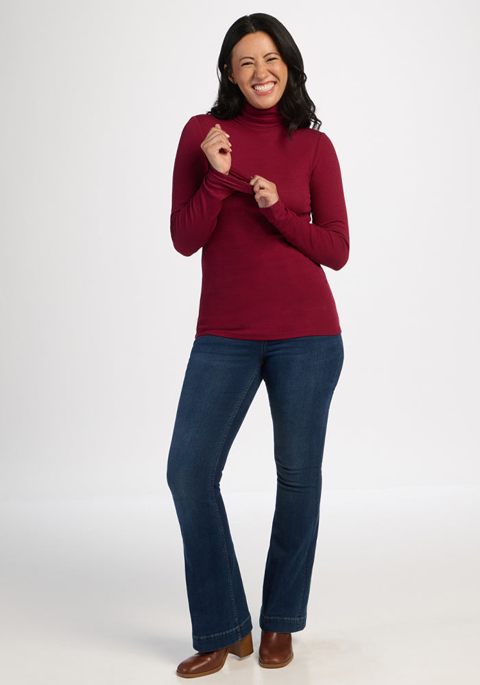 Woman in a Woolx Sage Ribbed Turtleneck - Beet Red, blue jeans, and brown boots smiling against a white background.