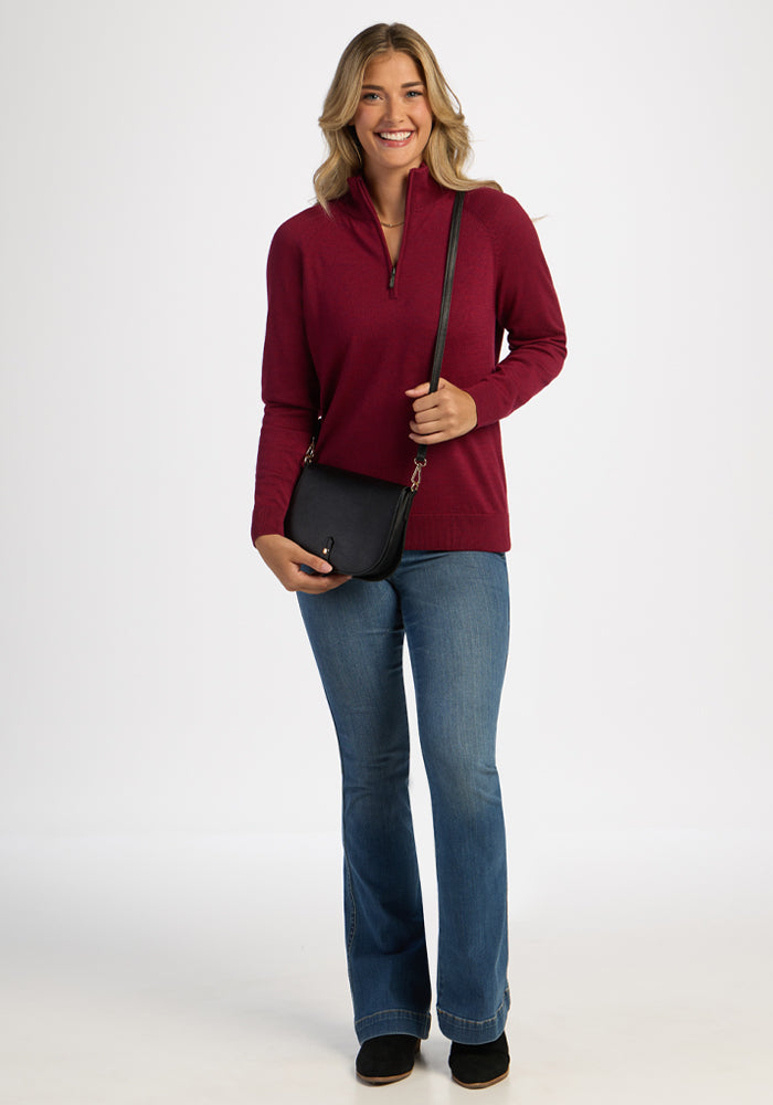 Smiling woman in Woolx Collins Half Zip Sweater - Beet Red, blue jeans, black shoes, holding a black crossbody purse.