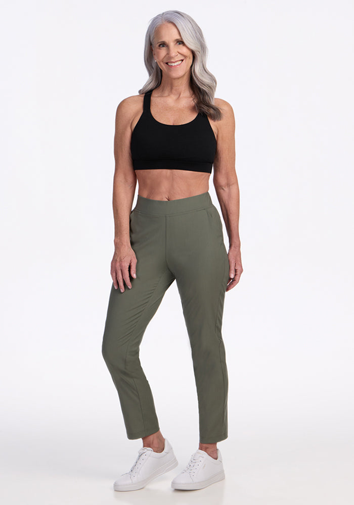 A smiling older woman with long gray hair wears a black sports bra, white sneakers, and Woolx Nicole Petite Ankle Pant in Sage Olive, standing confidently against a plain white background.