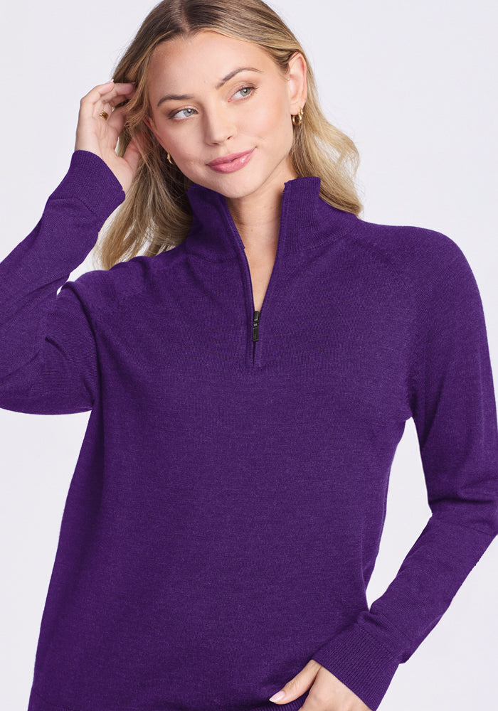 A woman with blonde hair and light skin smiles softly, touching her hair as she wears the Collins Half Zip Sweater in Plum Crush by Woolx against a light background.