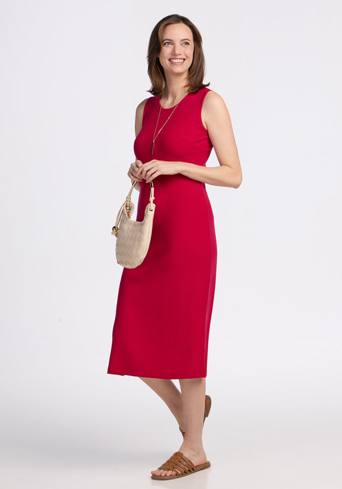 A woman smiles while wearing the Woolx Iris Ribbed Dress in Poppy Spark, paired with brown sandals, a simple necklace, and holding a small beige purse. She stands against a plain white background.