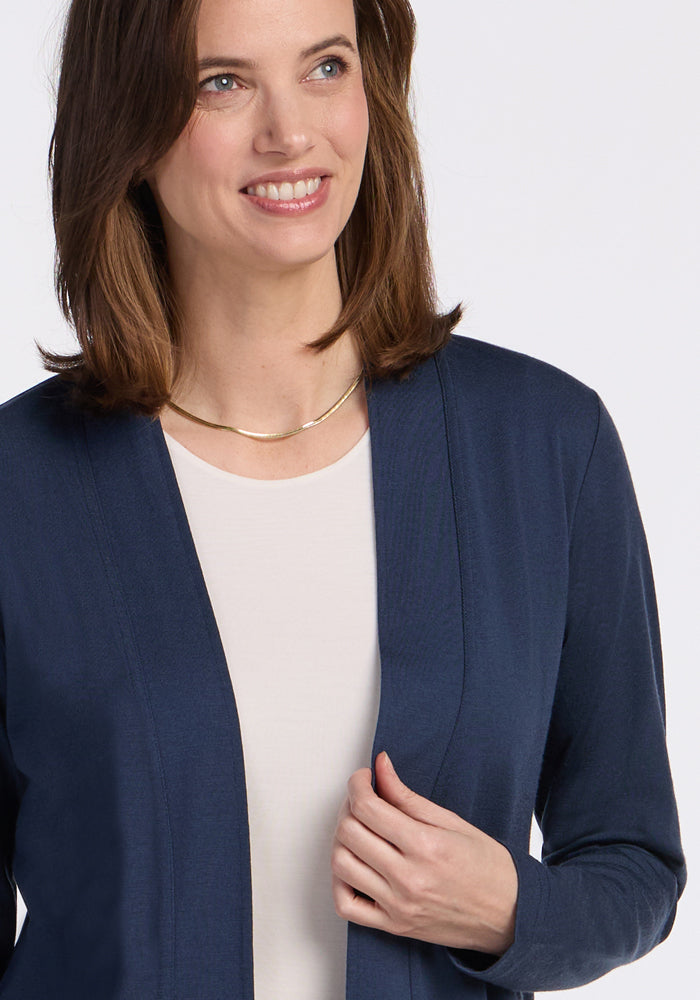 A woman with shoulder-length brown hair smiles, wearing the Woolx Paisley Cardigan in Deep Navy over a light top and a gold necklace, holding one side of the cardigan.