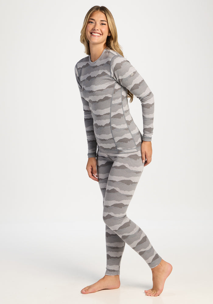 Woman wears Woolx Maddie Baselayer Top - High Peaks, standing barefoot and smiling against a white background.