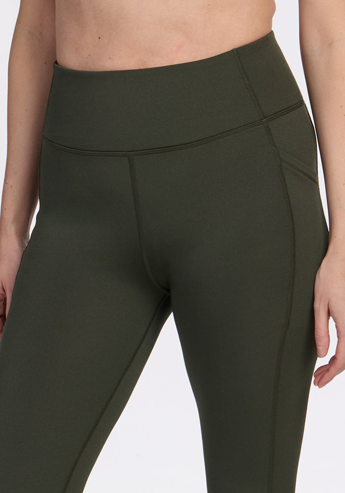 A person wears Woolx Merino Flex® McKenna Capris in Everleaf, high-waisted and dark green, shown from mid-torso to mid-thigh with one arm relaxed by their side against a plain background.
