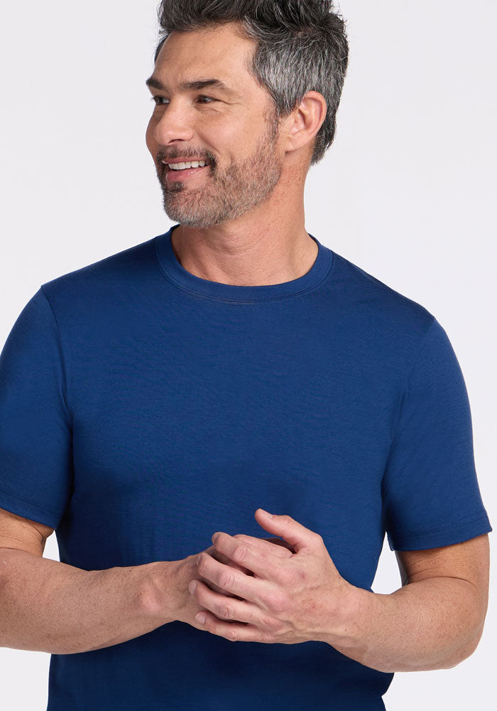 A man with short dark hair and a salt-and-pepper beard smiles and looks to the side, wearing the Woolx Endurance Tee in Estate Blue with his hands clasped in front of him. The background is white.