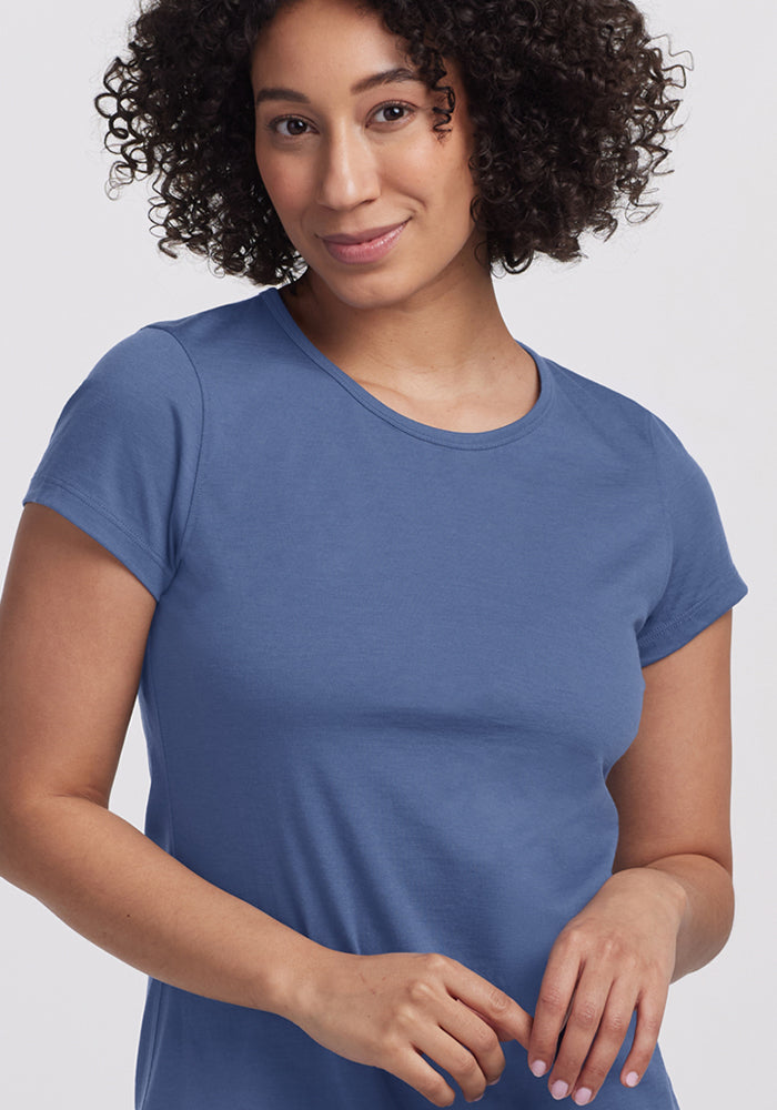 A woman with curly hair, wearing the Woolx Addie Crew Neck - Skyline short-sleeved shirt, smiles softly against a plain light background.