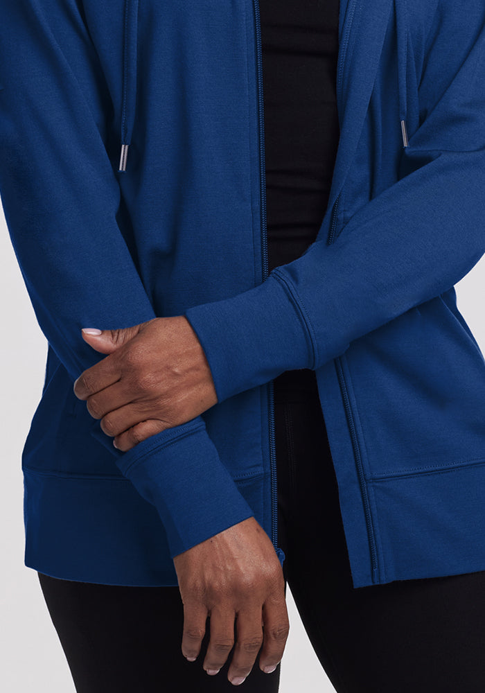 A person wearing the Woolx Ryann Hoodie in Estate Blue crosses one arm over, gently holding their opposite forearm. This travel essential features long sleeves with thumbholes and is paired with black pants against a plain background.