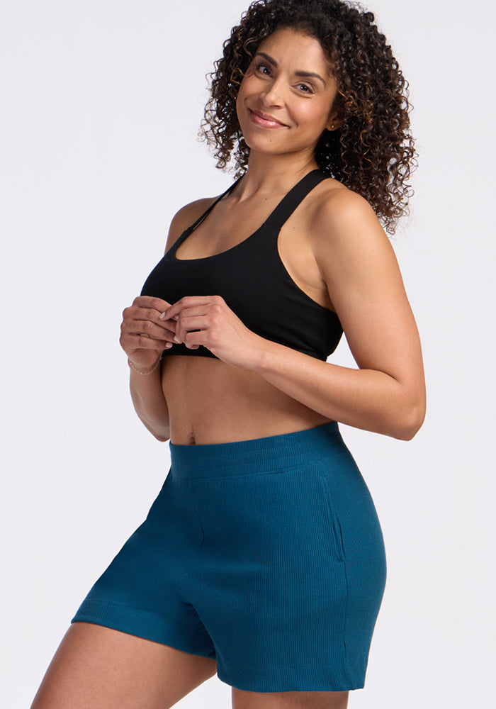 A woman with curly hair smiles while wearing a black sports bra and Woolx Maisey Ribbed Shorts in Moroccan Blue, standing against a plain white background.