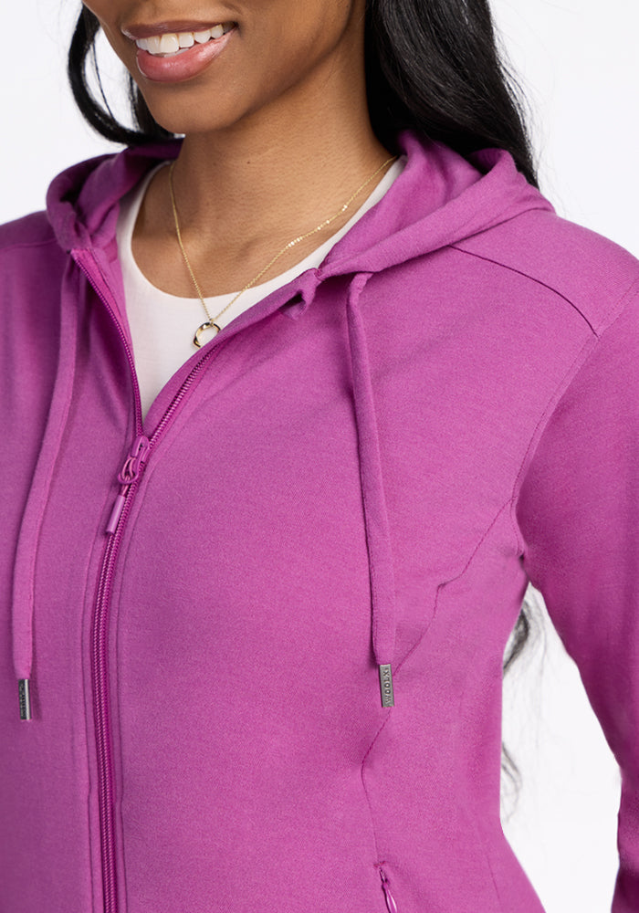 A woman smiles, wearing the Woolx Ryann Hoodie in Purple Wine over a white top with a gold necklace—visible from shoulders to mid-torso. This stylish, moisture-wicking layer is an ideal travel essential.