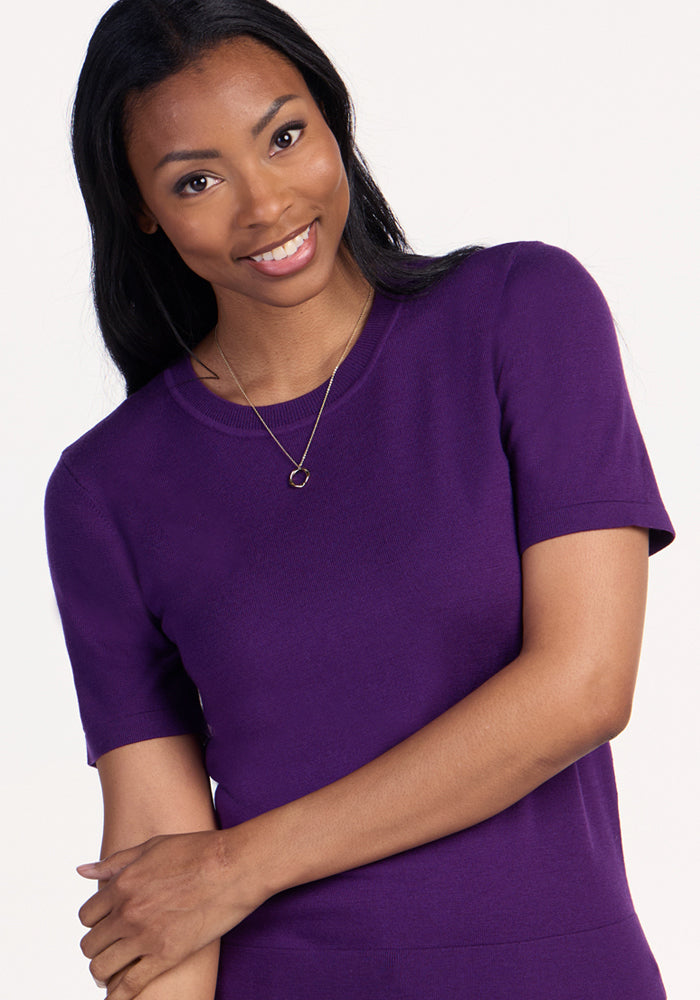 A woman with long dark hair, wearing the Woolx Anna Crew Sweater in Plum Crush and a simple necklace, smiles warmly at the camera against a plain light background.