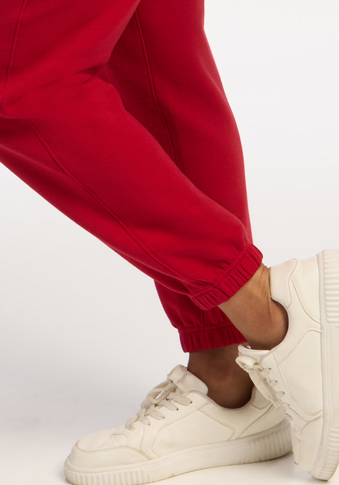 Model in Woolx Parker Sweatpants - Maraschino Cherry with white sneakers, one leg bent, on a plain white background.