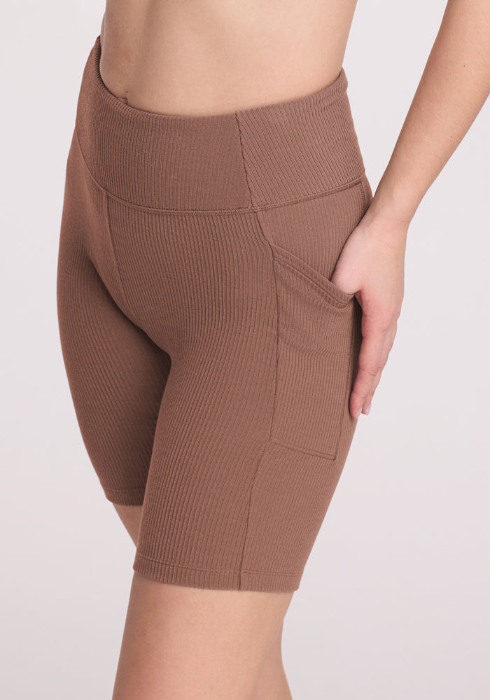 A person wearing Woolx Miley Bike Shorts in Sienna Kiss, featuring a high-waisted ribbed design and breathable fabric, with one hand in the pocket, is shown from waist to mid-thigh against a light backdrop.