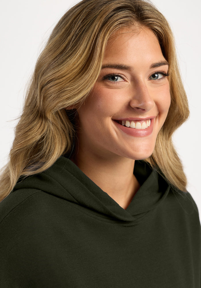 A smiling woman wearing a dark green Genevieve Hoodie - Duffel Bag made of temperature-regulating merino wool.