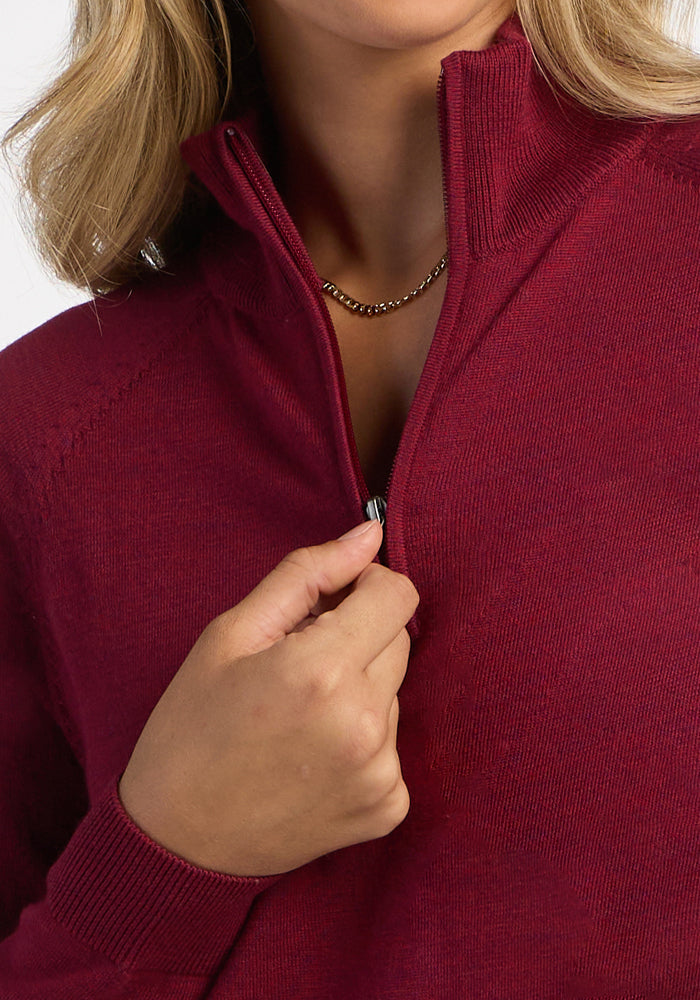 A person zips up the Woolx Collins Half Zip Sweater in Beet Red, wearing a gold chain and showing face and hand.