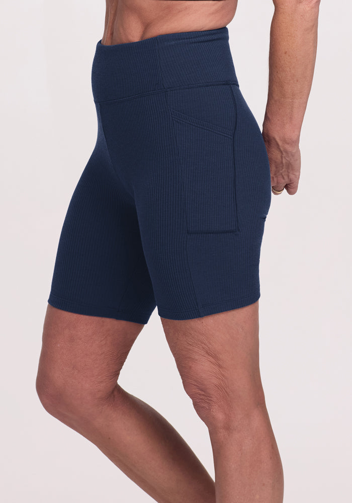 A person stands sideways against a light background, wearing Woolx Miley Bike Shorts in Deep Navy, featuring a high waist, ribbed texture, and side pockets.