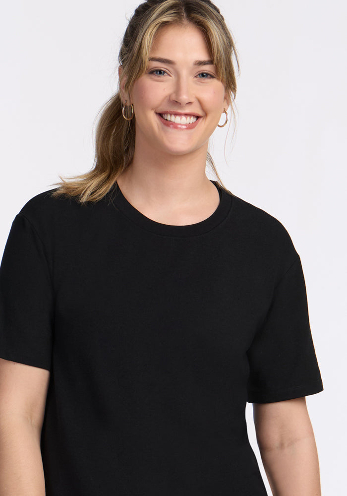 A young woman with light brown hair in a ponytail, wearing hoop earrings and the Woolx Audrey Tee in black, smiles at the camera against a plain white background.