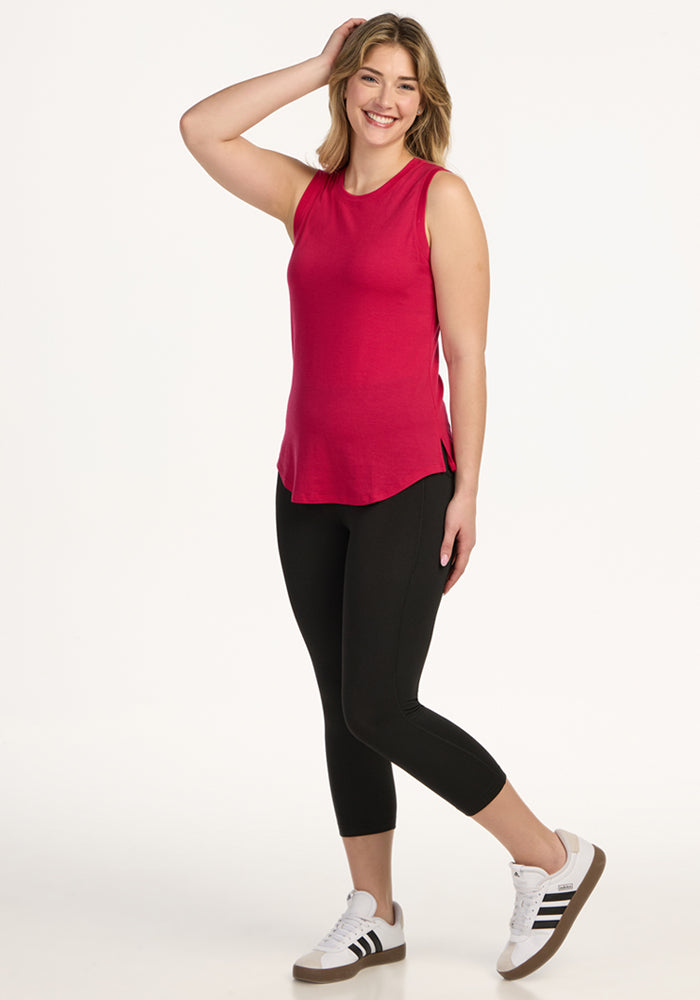 A smiling woman with shoulder-length blonde hair wears the Woolx Bella Tunic Top in Poppy Spark, black leggings, and white sneakers with black stripes. She stands with one hand touching her head against a plain white background.
