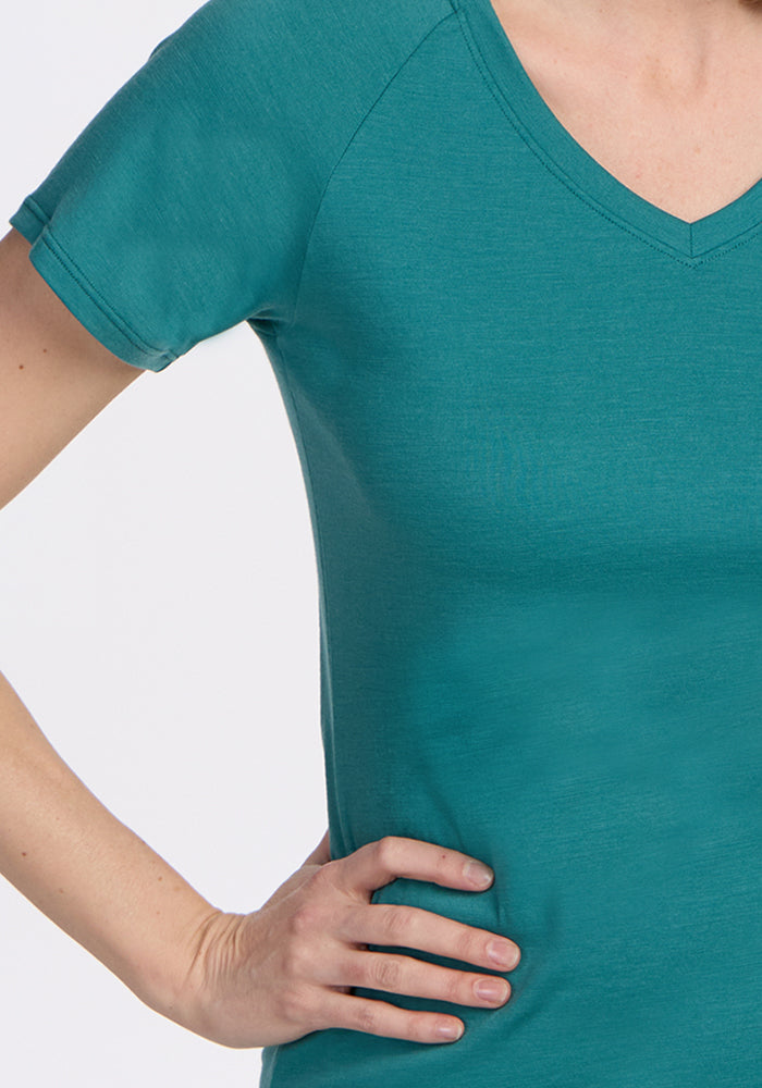A person models the Woolx Mia V Neck - Surfside, a teal women's Merino wool tee, standing with one hand on their hip against a plain white background. The image shows only the upper torso and part of the arm.