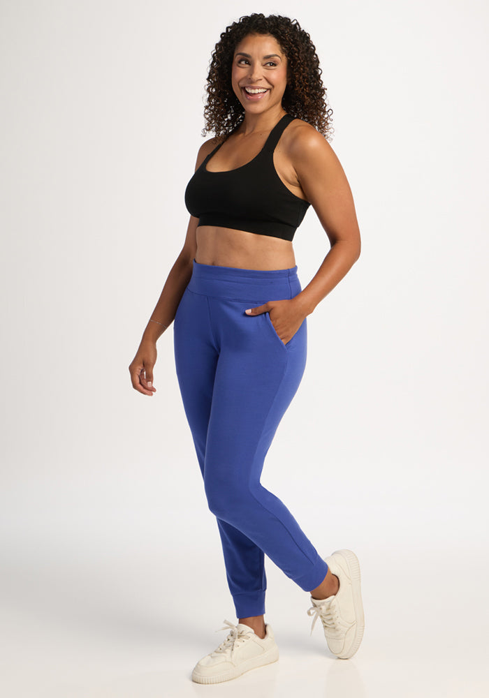 Woman in Woolx Madden Joggers - Royal Blue and black sports bra, smiling with hands in pockets on white background.