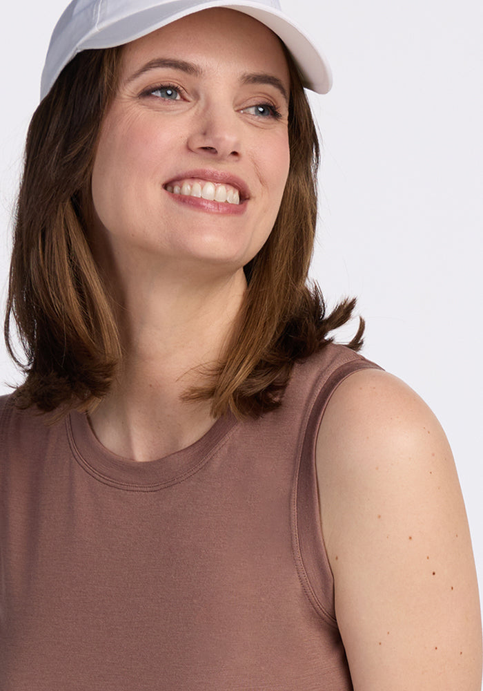 A smiling woman with shoulder-length brown hair wears the Woolx Bella Tunic Top in Sienna Kiss, paired with a white cap. She is looking slightly to the side against a plain light background.