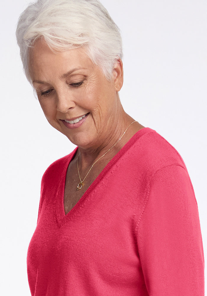 Smiling older woman in Woolx Francesca Sweater—Vivid Rose, gold necklace, looking down with short white hair.