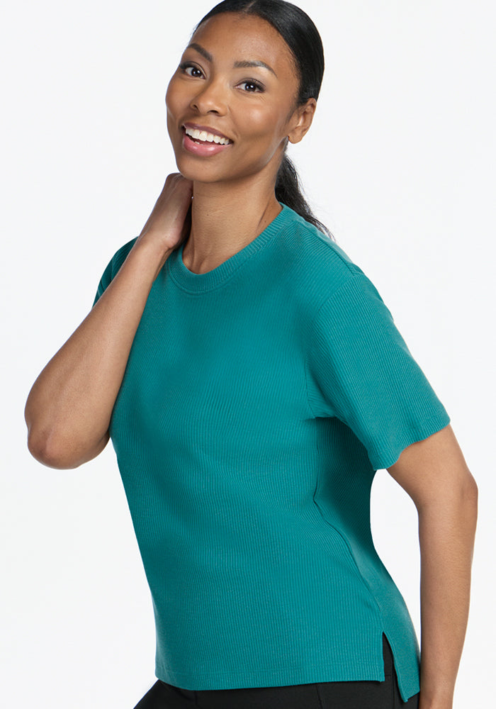 A woman with long dark hair smiles while wearing the Woolx Delia Ribbed Short Sleeve - Surfside and black pants, posing with one hand behind her neck against a plain white background.