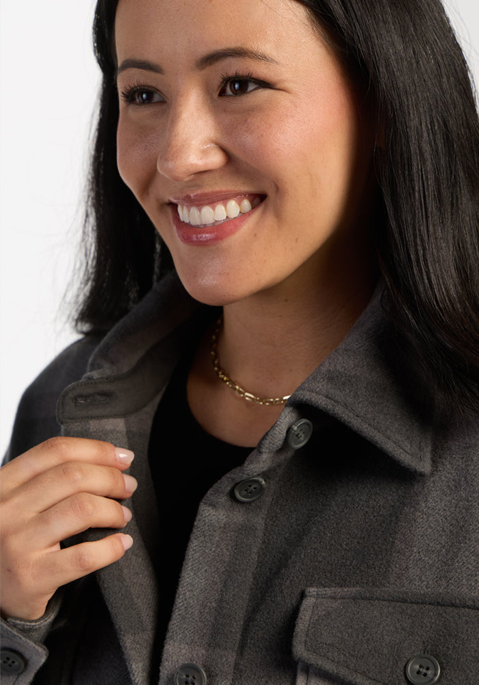 Woman smiling, wearing the Woolx Sawyer Shacket in Volcanic Ash Plaid, with a black top and gold chain necklace.
