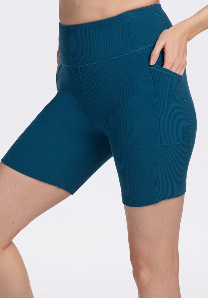 A person wearing Woolx Miley Bike Shorts in Moroccan Blue, featuring a high-waisted ribbed design and side pocket, stands against a plain background with one hand in the pocket.