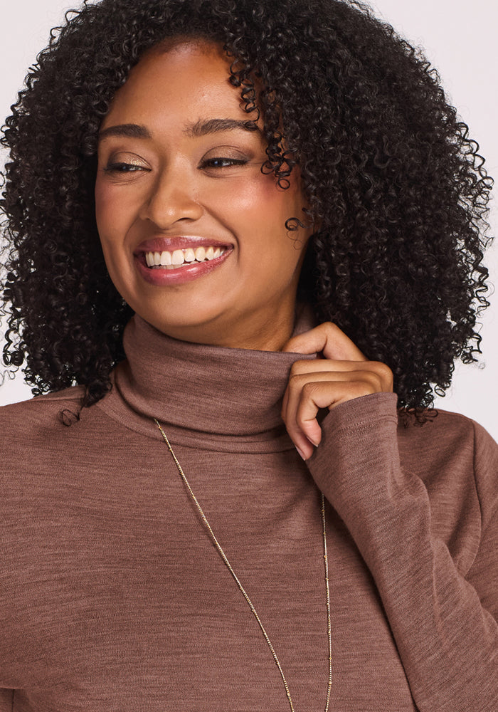 Smiling woman with curly hair wears the Woolx Peyton Turtleneck in Sable Heather and a long necklace, looking aside.