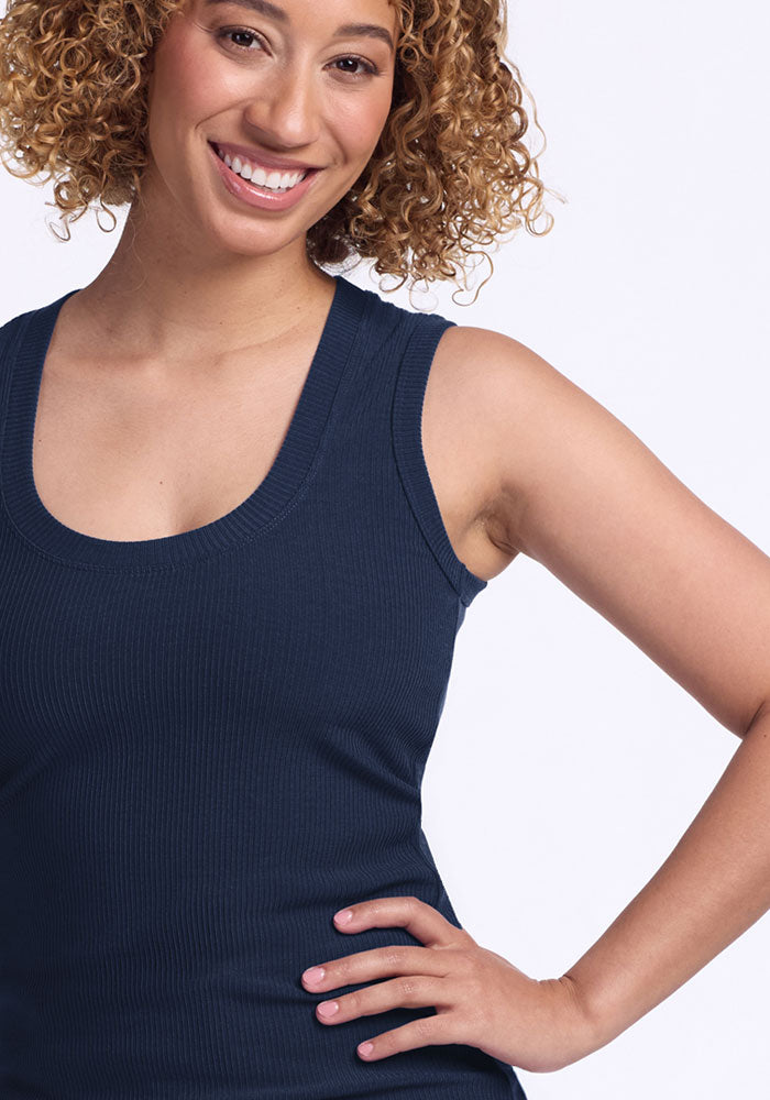 A smiling woman with curly hair wears the Woolx Sloane Ribbed Tank in Deep Navy. She stands with one hand on her hip against a plain white background.