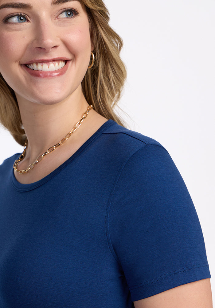 A woman with wavy blonde hair smiles while wearing the Woolx Liza Crew Neck Tee in Estate Blue, paired with a gold chain necklace and hoop earrings. She is shown from the shoulders up against a plain white background.