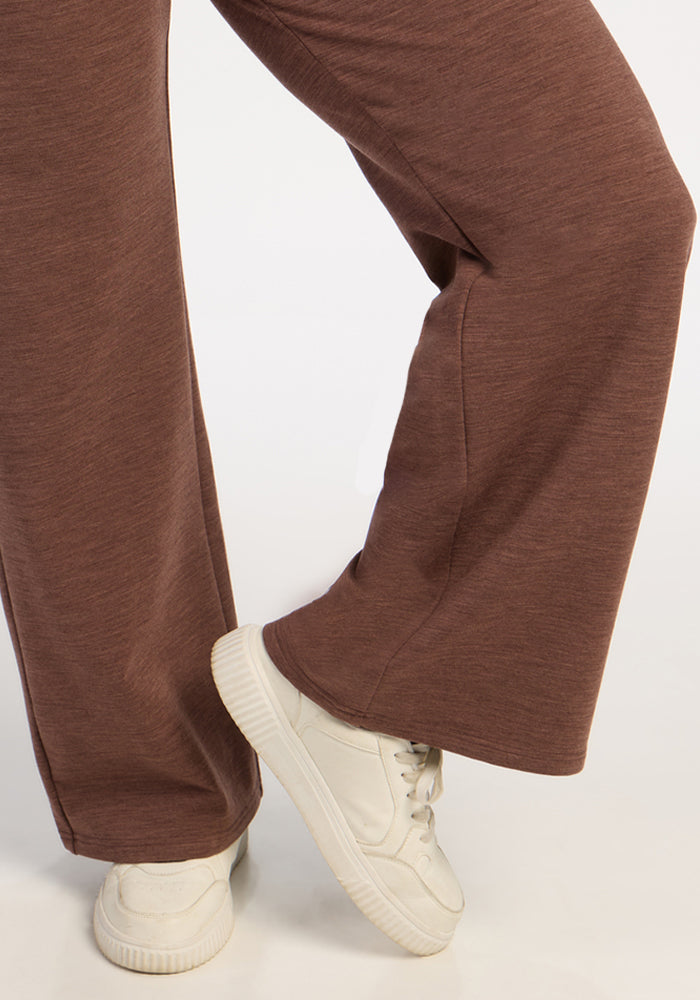 Individual in Woolx Eloise Wide Leg Pants - Sable Heather and white sneakers, standing with one leg slightly bent.