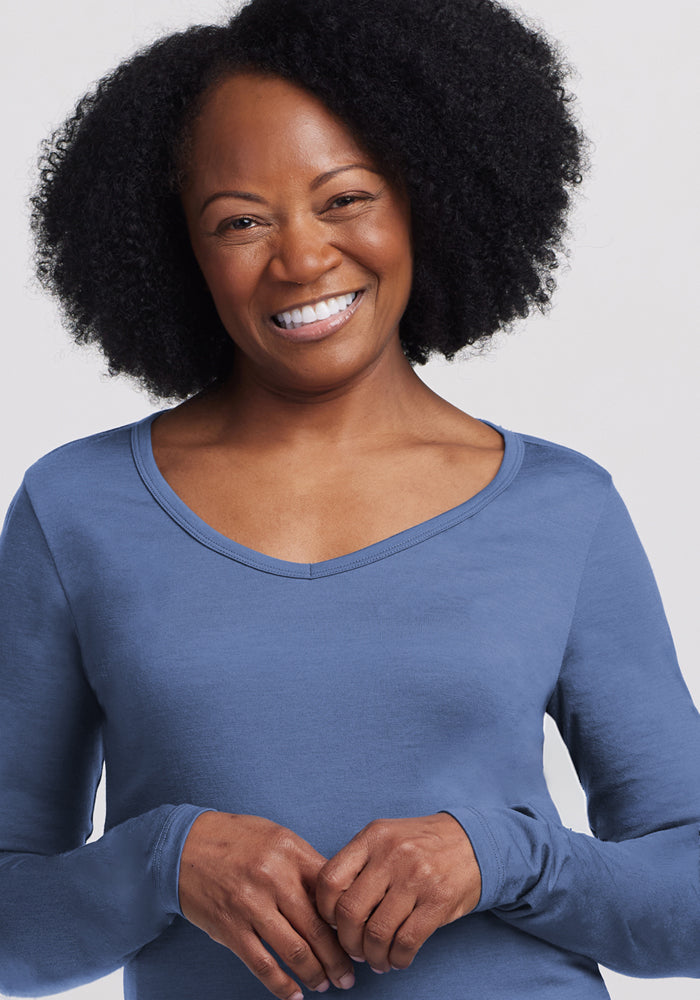 A smiling woman with curly natural hair wears the Woolx Layla V Neck Top in Skyline, a long-sleeved light blue Merino wool shirt, standing against a plain light background and looking at the camera with her hands gently clasped.