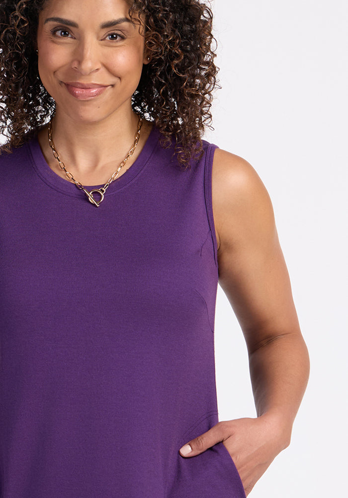 A woman with curly hair smiles at the camera, wearing the Woolx Cassie Dress in Plum Crush and a gold chain necklace with a heart-shaped pendant, with one hand in her pocket.