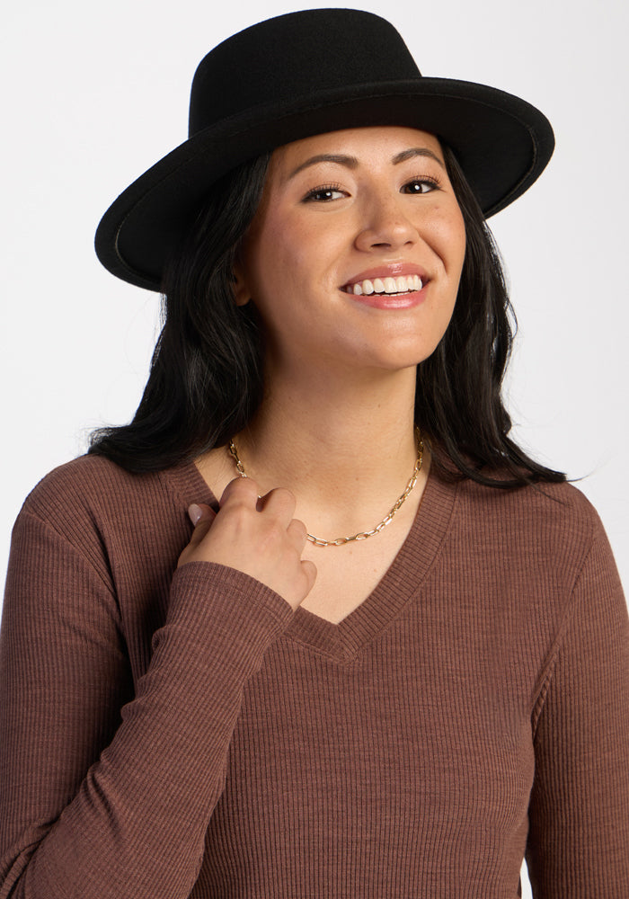 Smiling woman in a black hat and Woolx Evie Ribbed Tunic - Sable Heather, gold chain necklace, plain backdrop.
