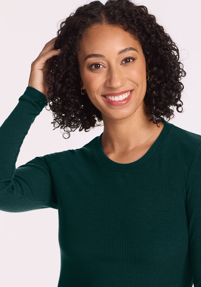 Smiling woman in Woolx Hadley Petite Crew - Deep Pine, touching her curls, stands against a plain background.