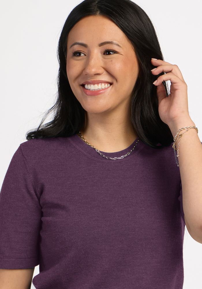 Woman in Woolx Anna Crew Sweater - Purple Iris smiles, looking away and touching her long dark hair, layered necklaces on.