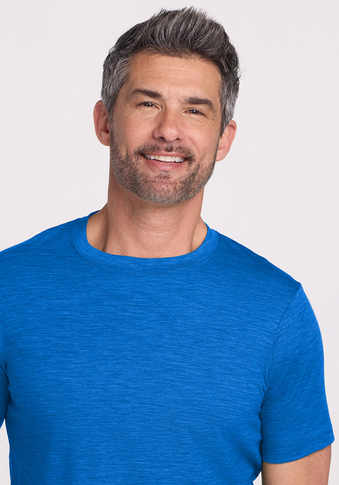 A smiling man with short gray hair and a trimmed beard wears the Woolx Endurance Tee in Glacial Blue, standing against a plain light background.