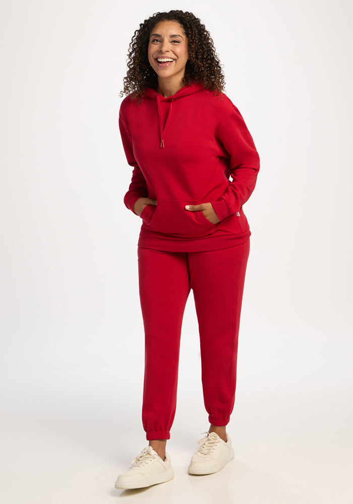 Woman in Woolx Avery Hoodie - Maraschino Cherry, matching joggers, smiles with hands in pockets against white background.